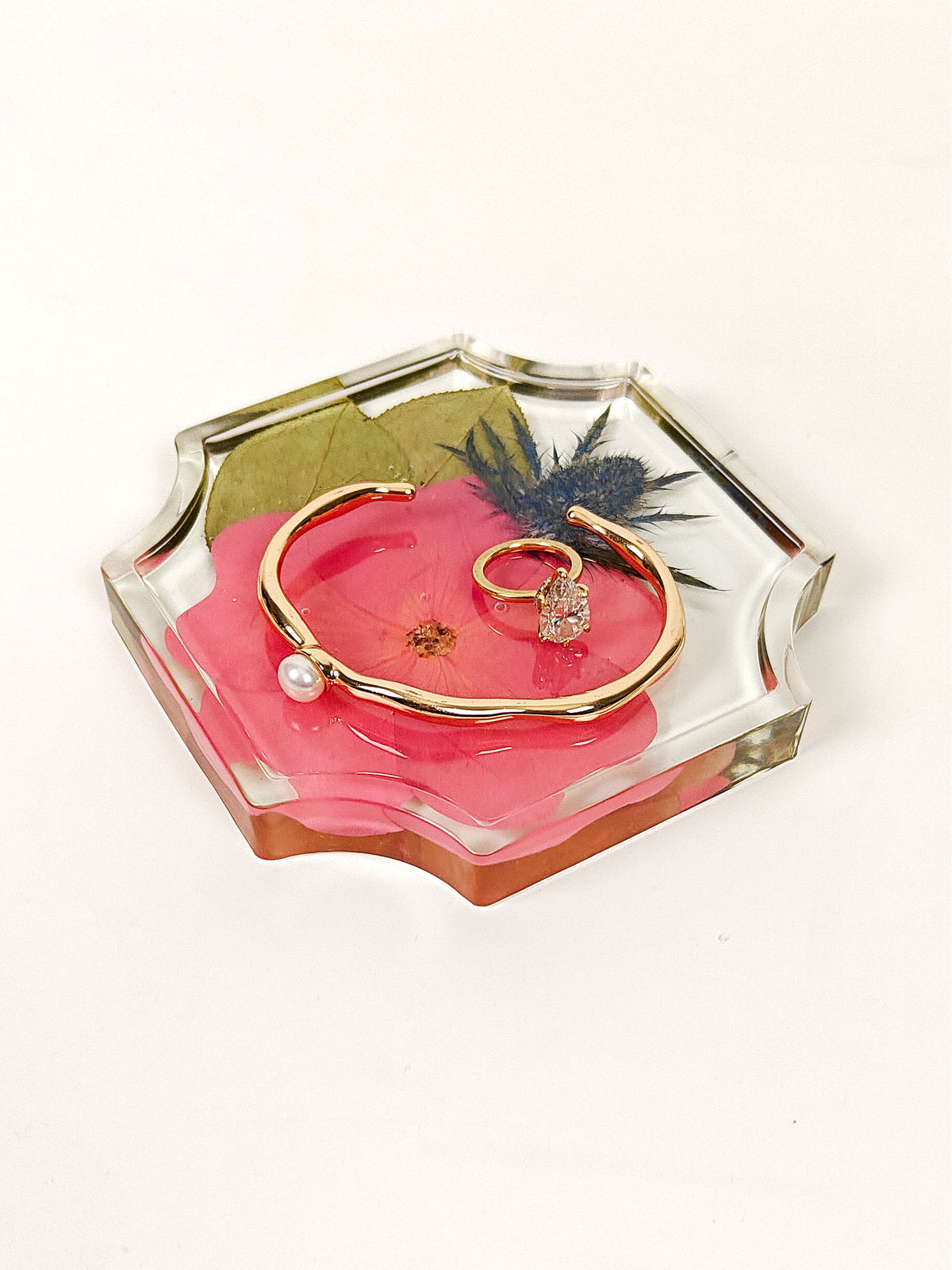 A gold-tone open bangle bracelet with a small pearl bead and a gold ring with a clear stone, displayed inside a clear acrylic hexagonal tray filled with pink flower petals and dried botanicals, on a white background.