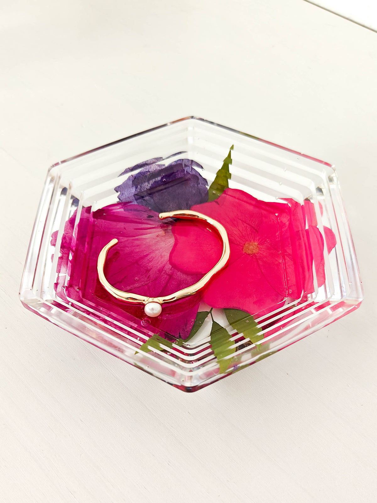 A gold open cuff bracelet with a small white pearl accent, displayed inside a clear hexagonal glass dish filled with bright pink and purple flower petals on a white surface.