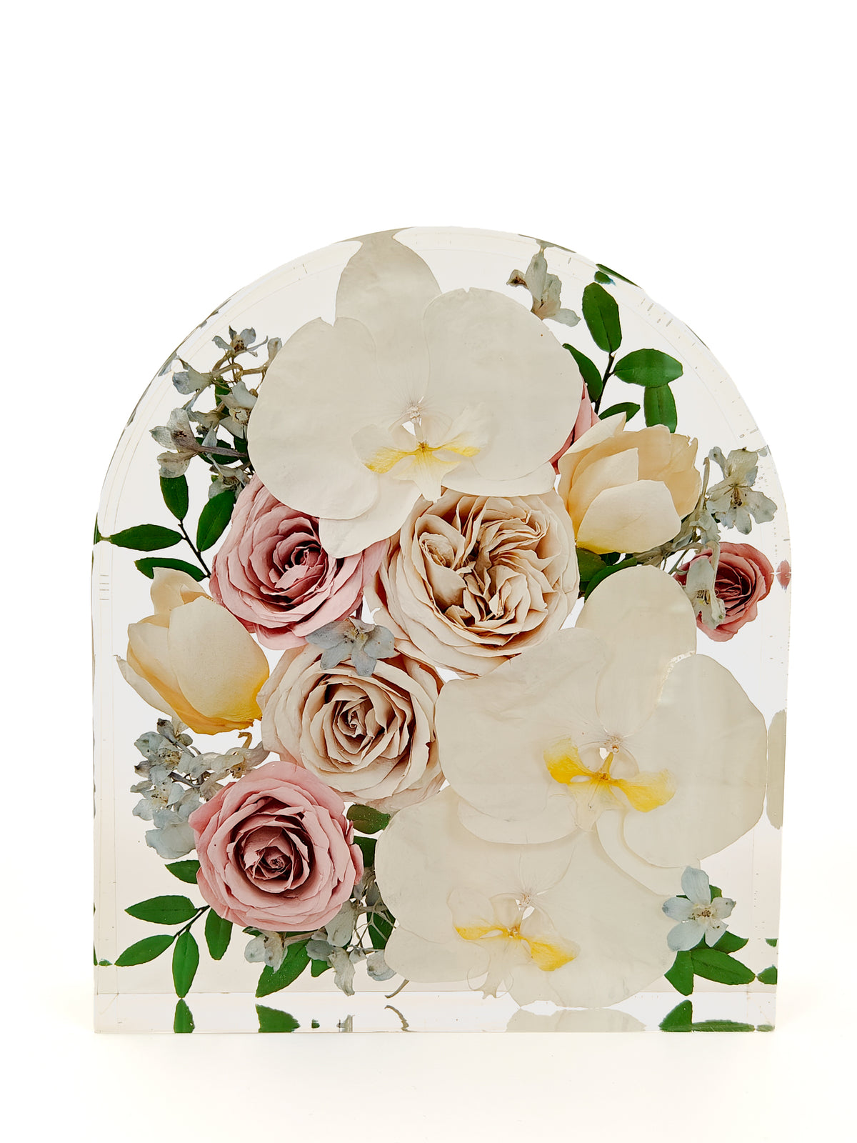 An arched clear resin plaque featuring a pressed floral arrangement with large white orchids, blush pink and cream roses, green leaves, and small pale blue flowers, photographed on a white background.