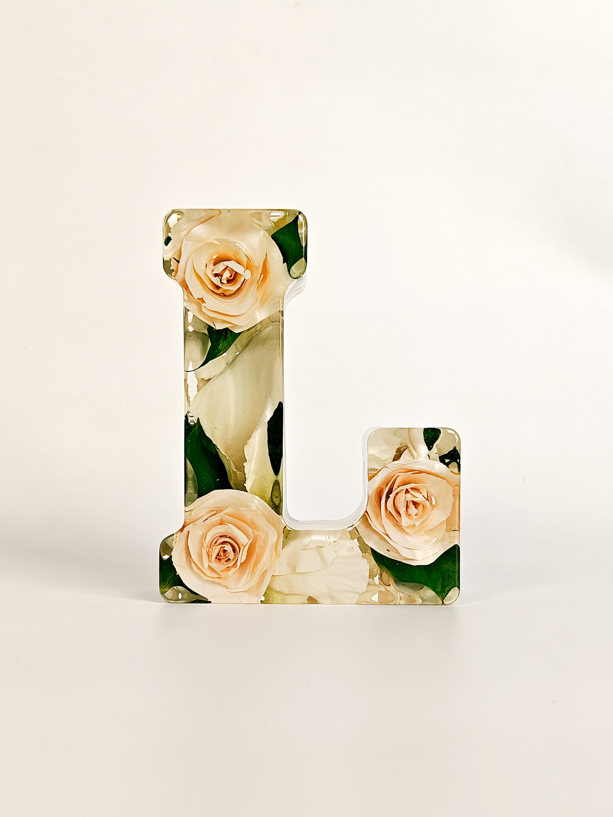 A decorative letter 'L' made of clear resin with a printed floral design of peach roses and green leaves, standing on a light beige surface against a white background.