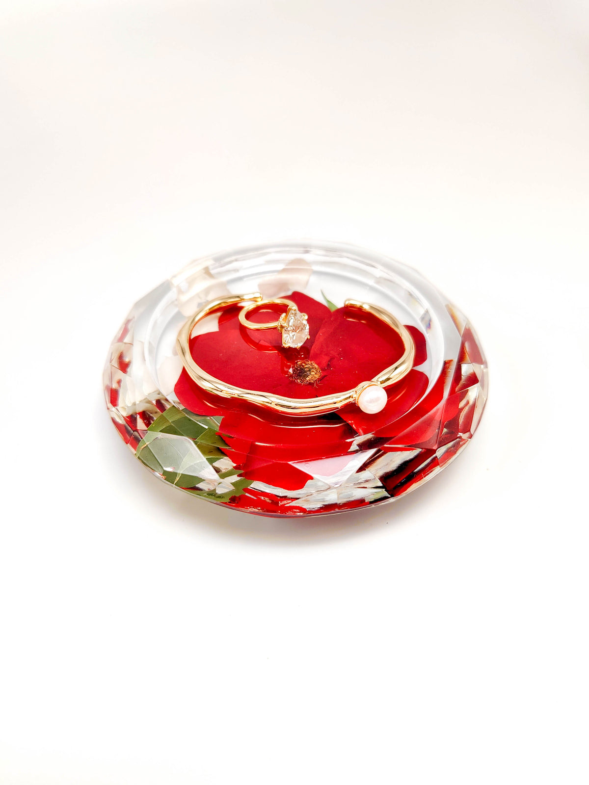 A heart-shaped clear resin keepsake box filled with red rose petals, green leaves, and a gold ring with a clear gemstone, displayed on a white background.