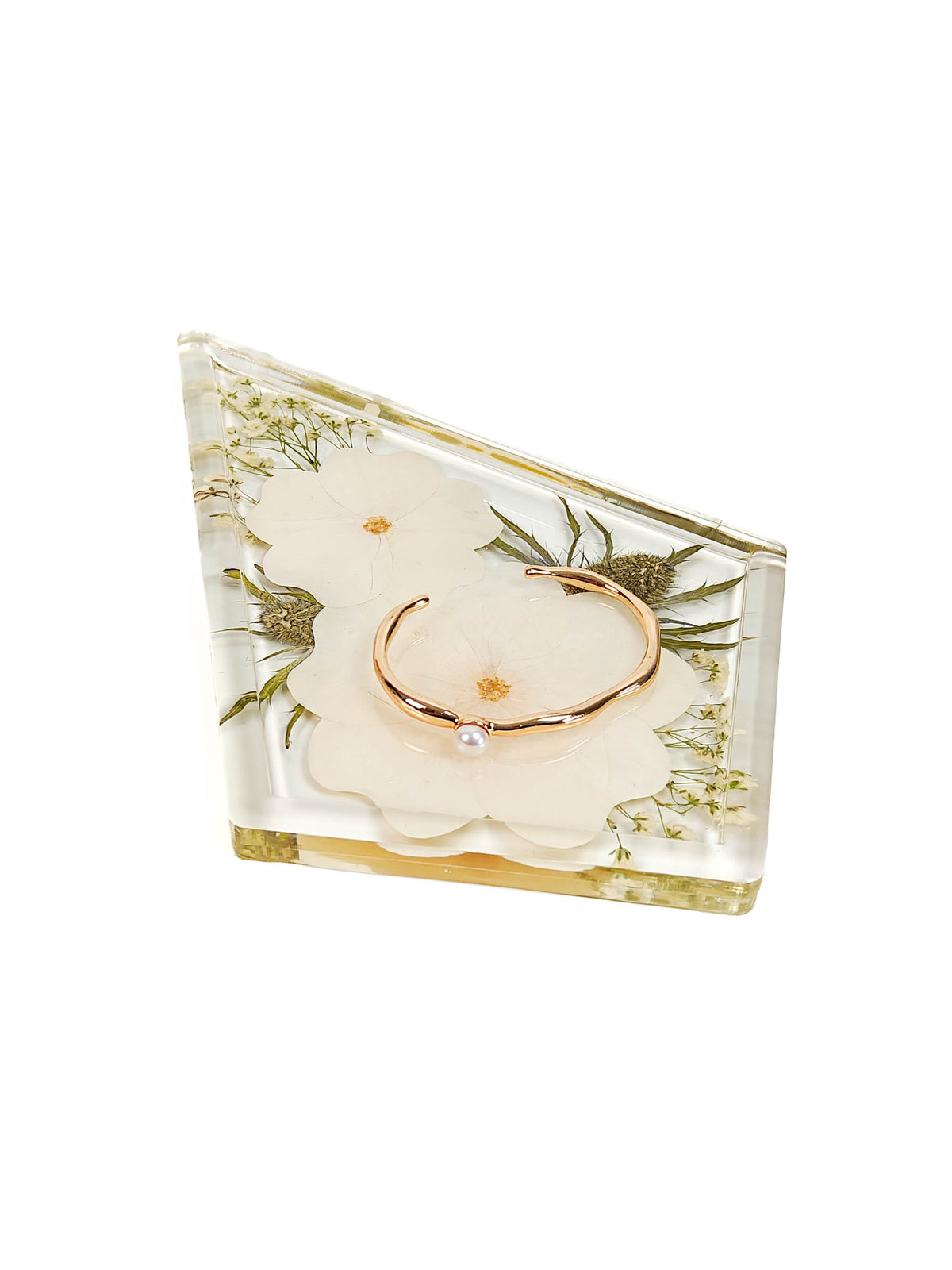 A triangular clear resin box containing dried white flowers and green leaves, with a rose gold ring resting inside on a white background.