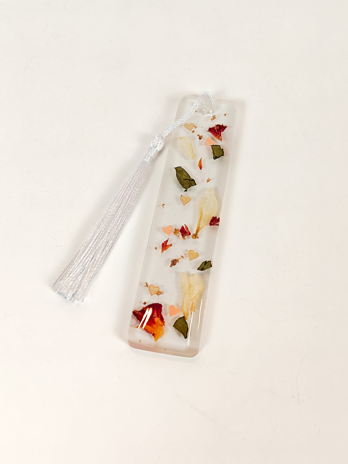 A clear rectangular resin bookmark with dried flower petals and leaves inside, finished with a white tassel, shown on a plain white background.