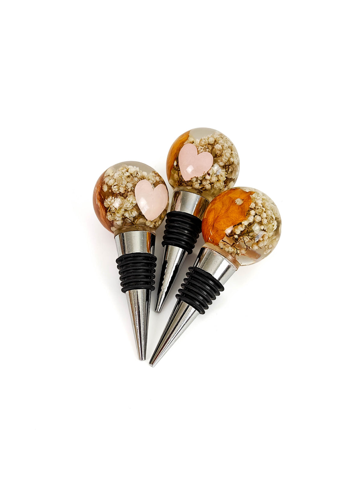 Three wine bottle stoppers with clear spherical tops filled with gold glitter and a small pink heart, attached to black rubber seals and silver metal points, on a white background.