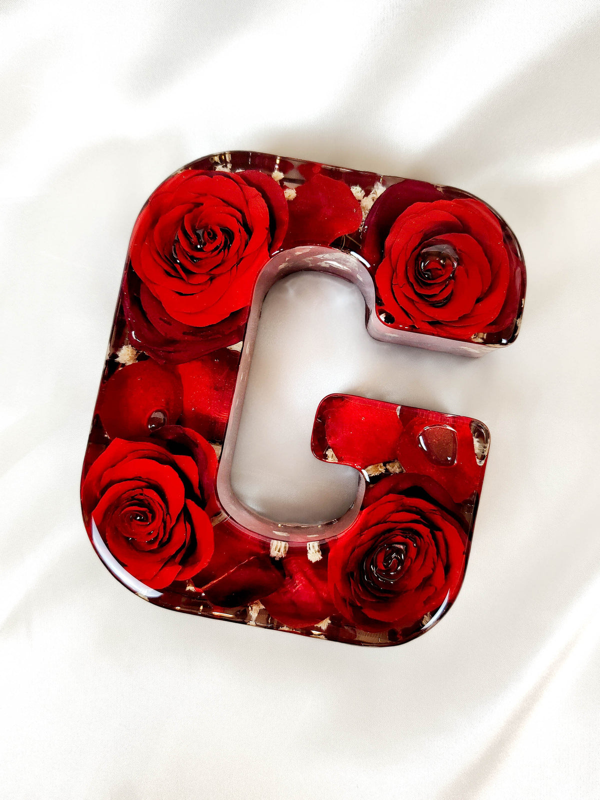 A decorative resin letter 'G' filled with red roses and gold leaf, photographed from above on a white fabric background.