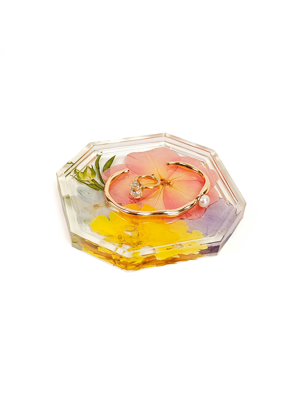 A clear resin keepsake box with a geometric octagonal shape, containing a pink hibiscus flower, yellow petals, and a gold ring, shown on a white background.