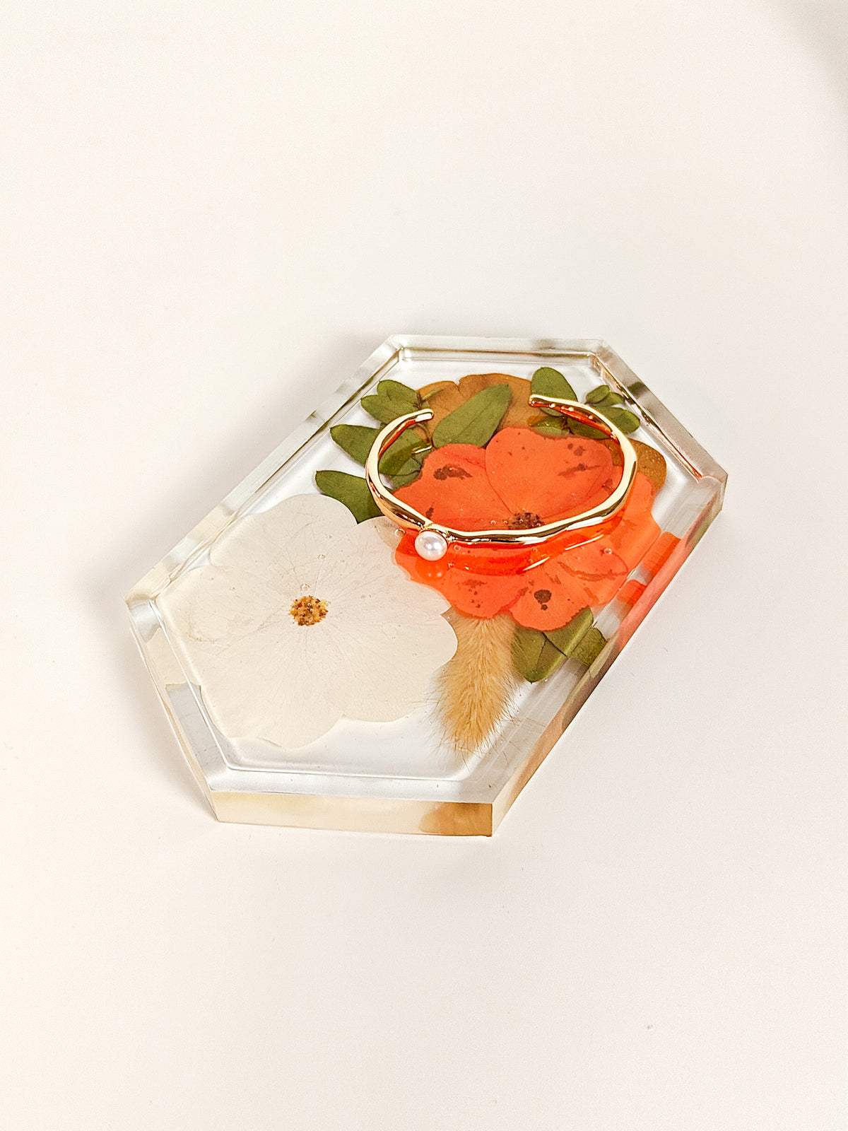 A clear acrylic hexagonal tray containing dried botanical elements, including a white flower, orange petals, green leaves, and a gold-toned bracelet, photographed on a clean off-white background.