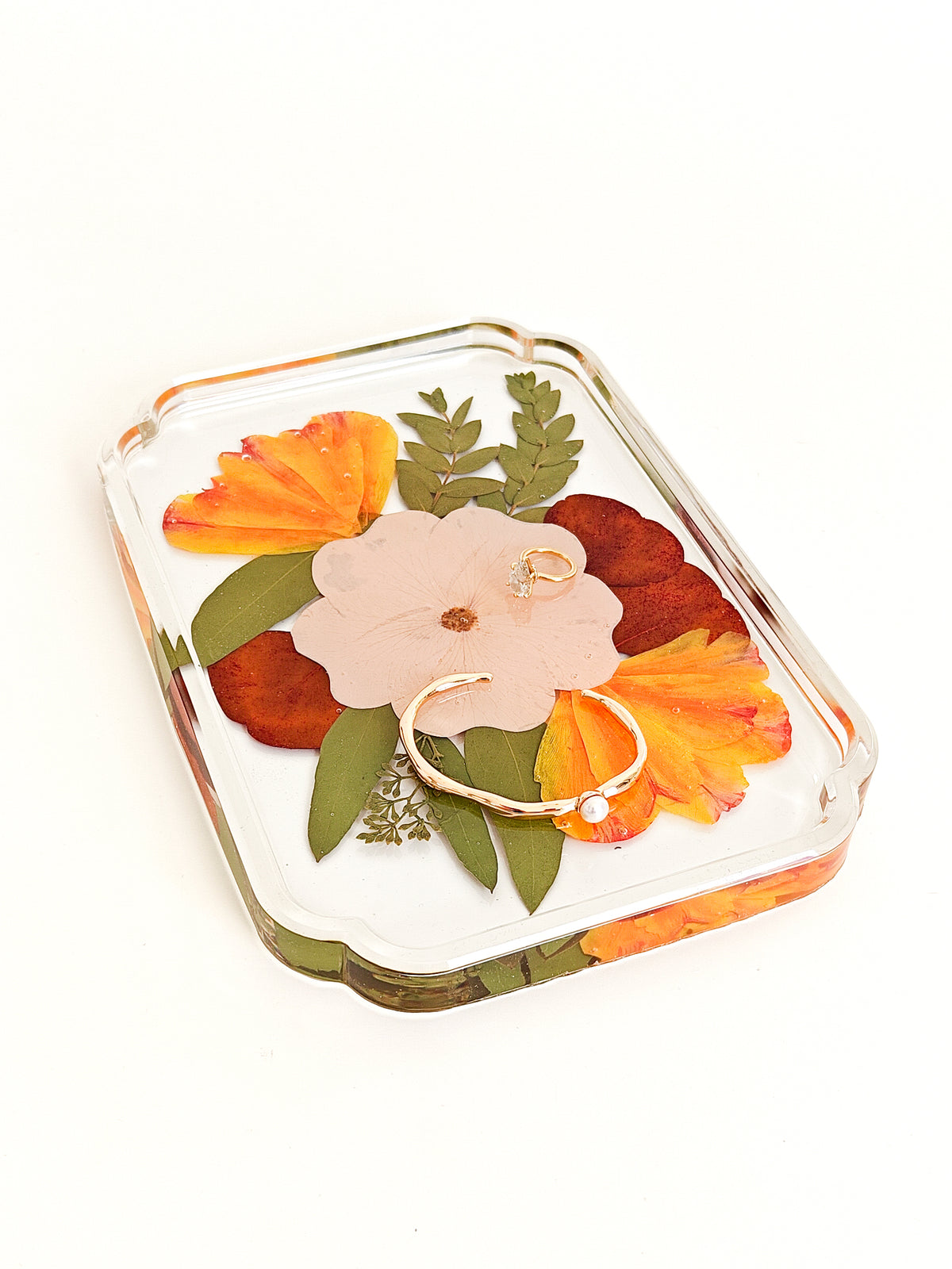 A clear acrylic jewelry tray with a scalloped edge, filled with pressed orange flowers, green leaves, and a pale pink flower, holding two gold rings, on a white background.
