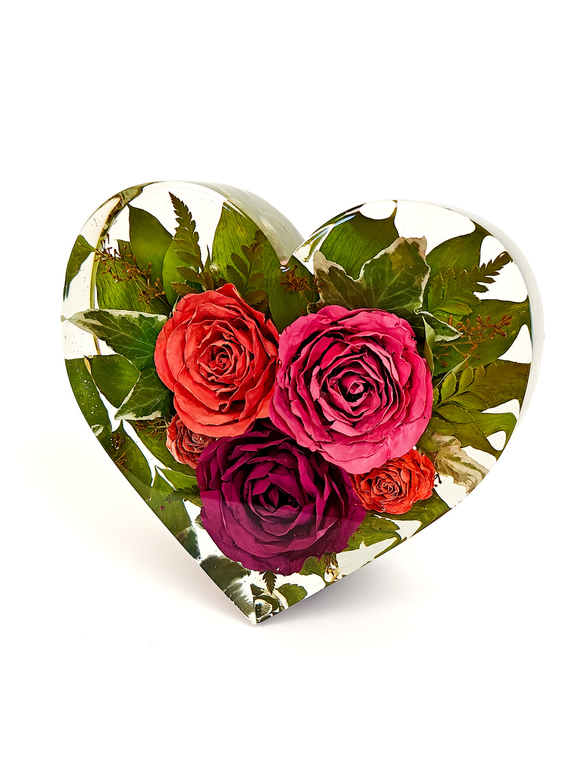 A heart-shaped decorative object featuring a preserved floral arrangement with red, pink, and purple roses and green leaves inside clear resin, shown on a white background.