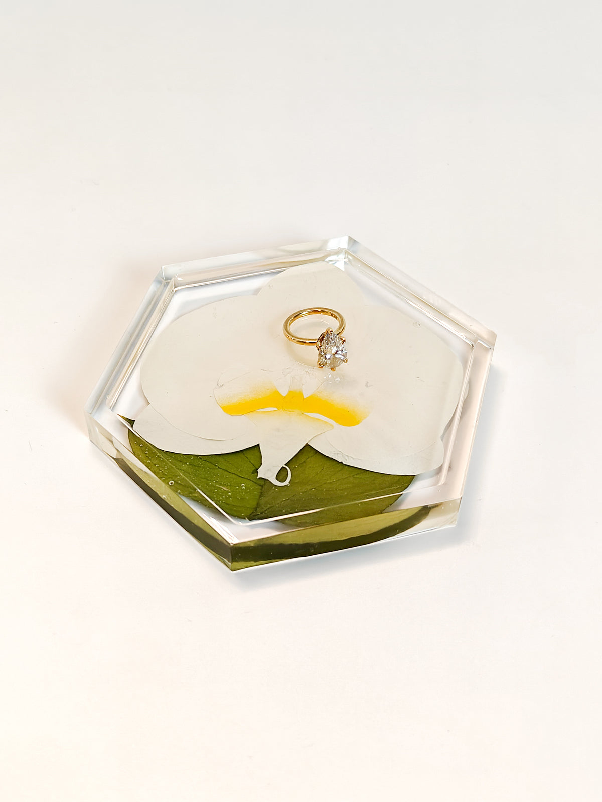 A hexagonal clear resin ring box containing a white orchid flower, green leaf, and yellow accent, with a gold ring displayed inside, set against a plain white background.