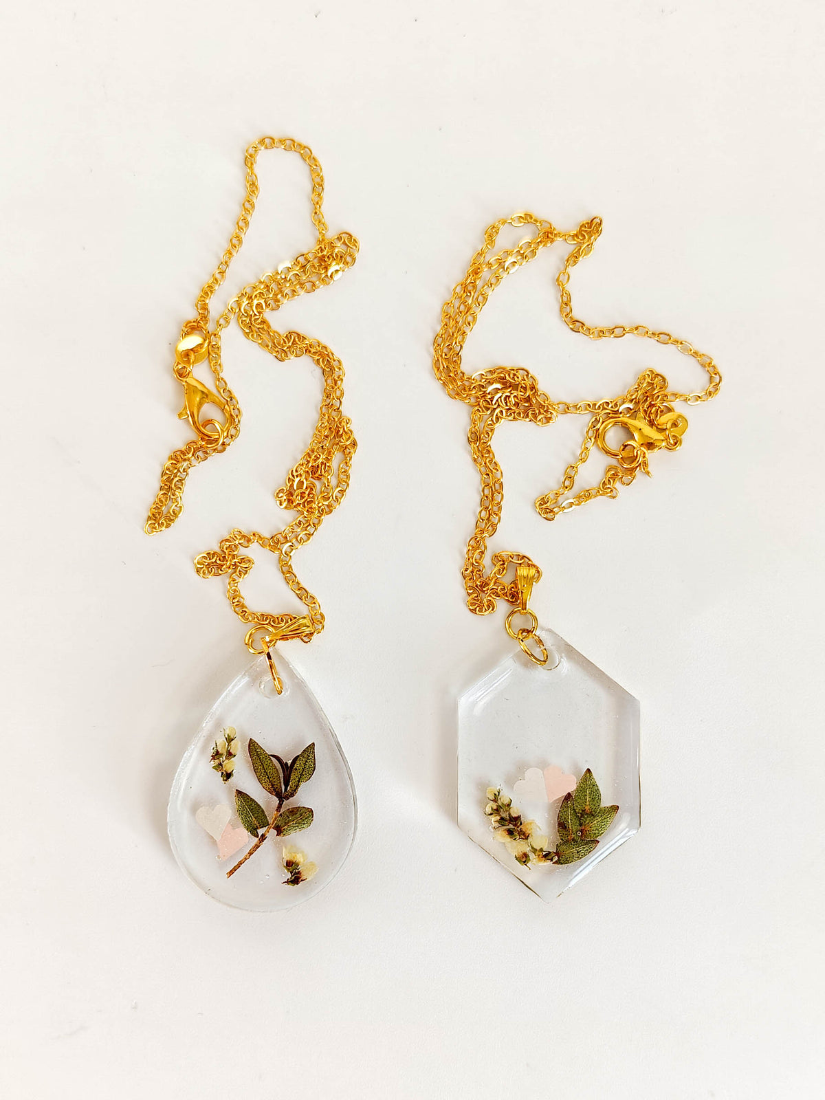 Two gold chain necklaces with clear resin pendants containing pressed flowers and leaves, displayed on a white background.