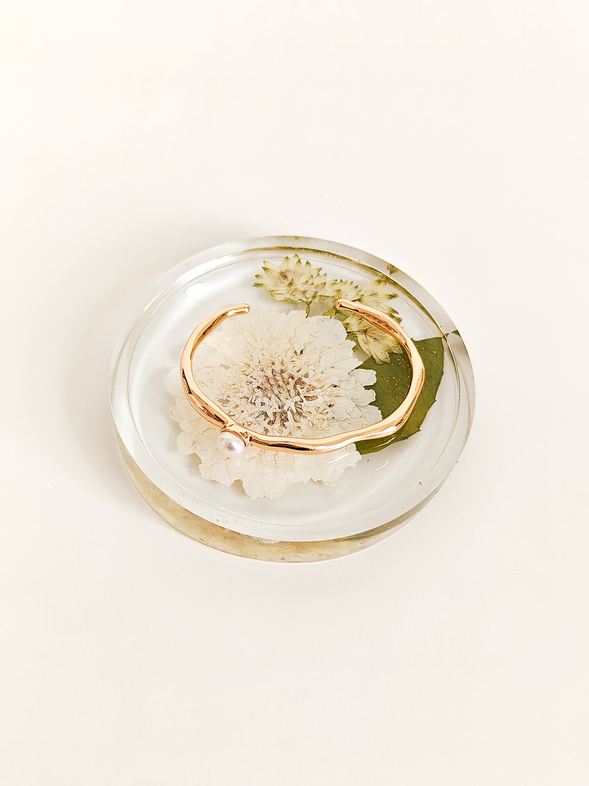 A thin open cuff bracelet in a warm rose gold tone, displayed on a small round clear resin tray with dried flowers and a green leaf, set against a clean off-white background.