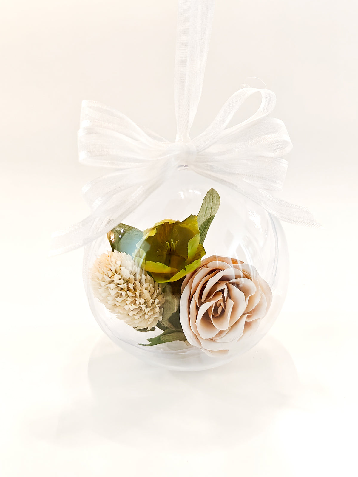 A clear glass hanging ornament filled with dried flowers, including a pale peach rose and a beige dahlia, with green leaves and a white ribbon bow at the top, set against a clean white background.