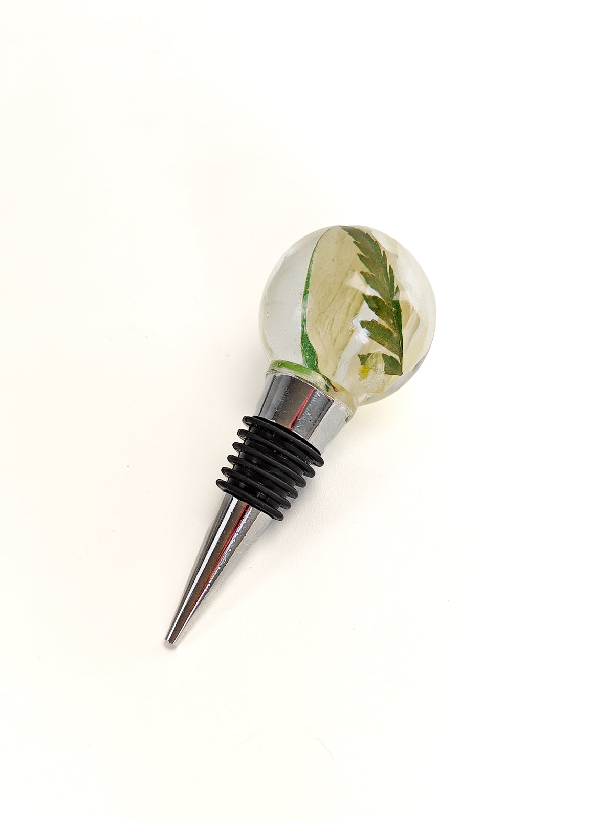 A clear glass wine stopper with a silver metal spike and a black rubber grommet, featuring a decorative top containing dried green leaves and a pale yellow flower.