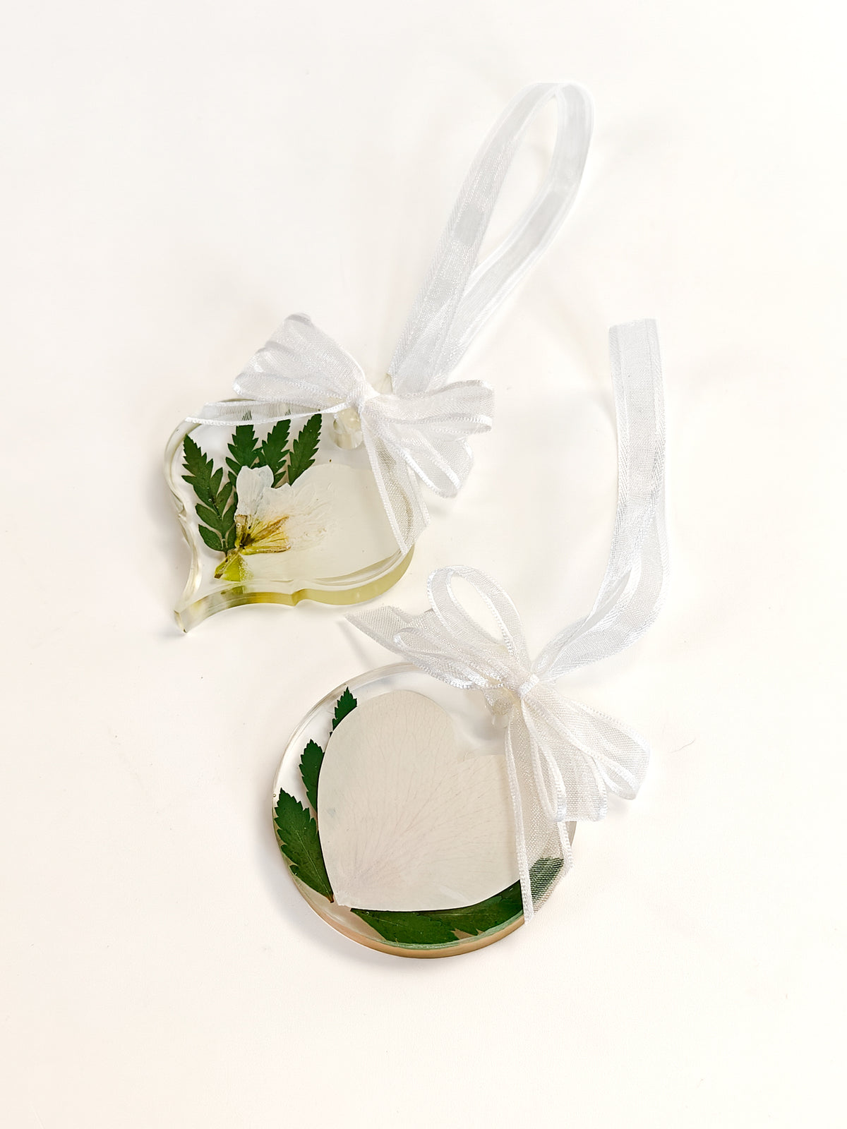 Two resin hanging ornaments on a white background, one irregularly shaped with a green leaf and yellow flower, and one round with a white feather and green leaf, both tied with white ribbon.