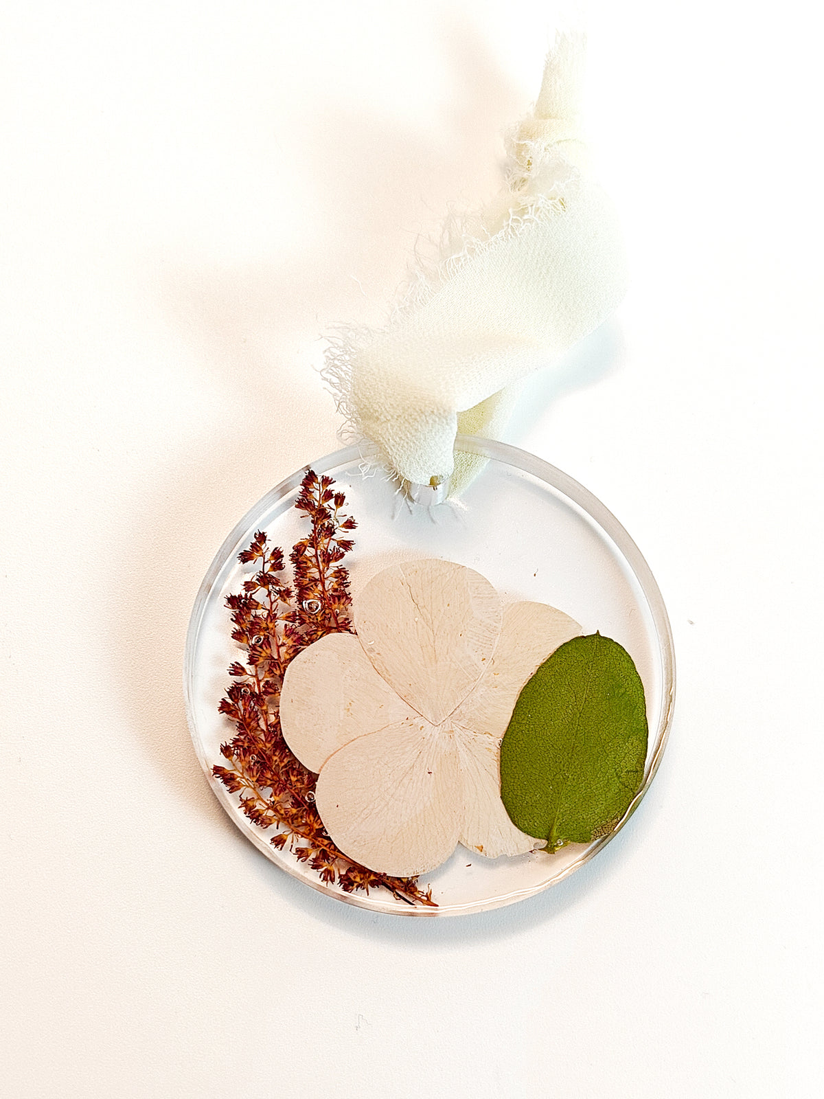 Vyvy Pham A circular clear resin keepsake or ornament containing pressed botanical elements, including a pale flower, a green leaf, and reddish dried flowers, with a white fabric loop at the top, photographed on a white background.