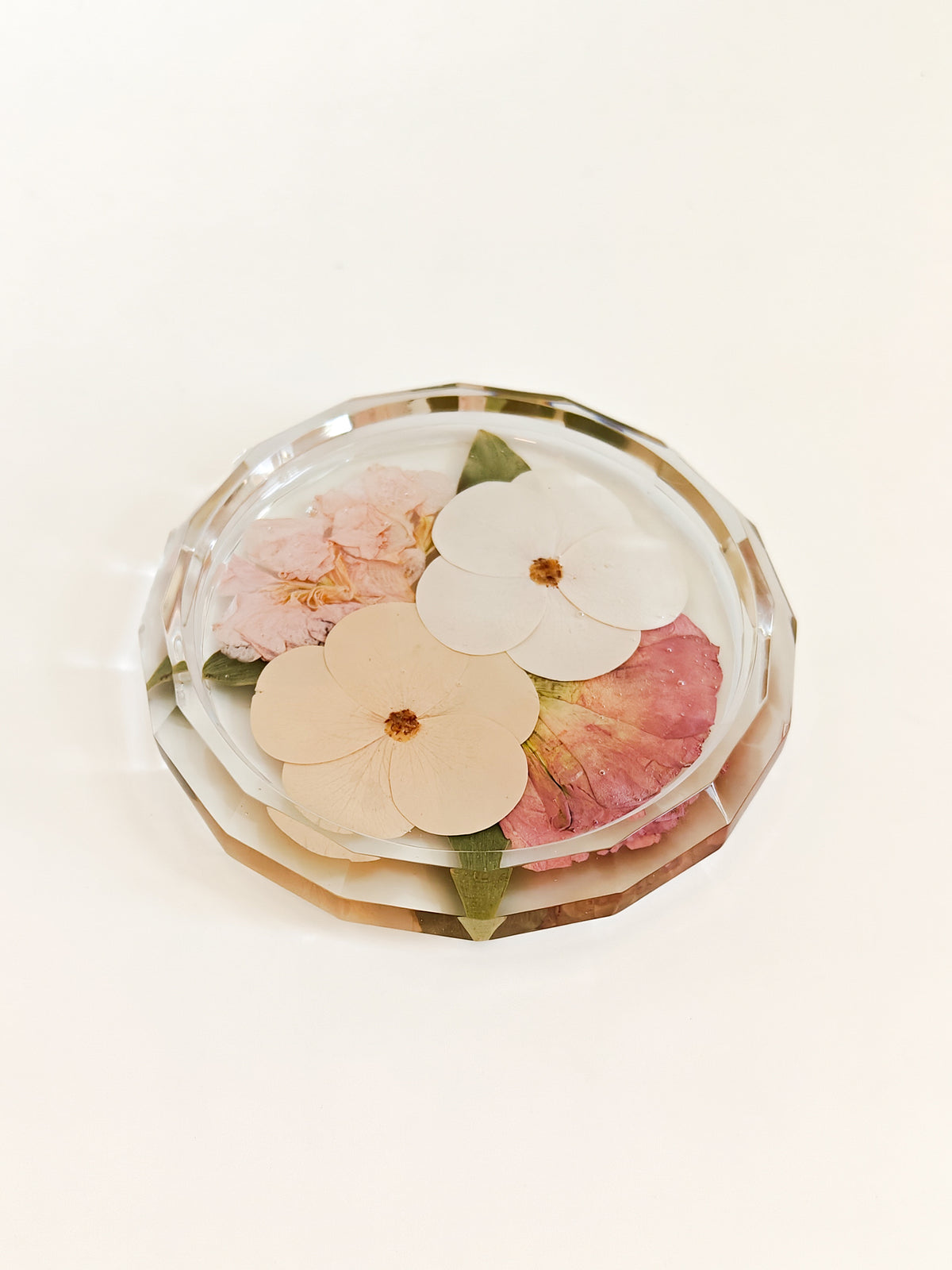 A round clear resin or glass dish with a faceted edge, containing pressed flowers in soft pink, cream, and peach tones with small green leaves, photographed on a clean white background.