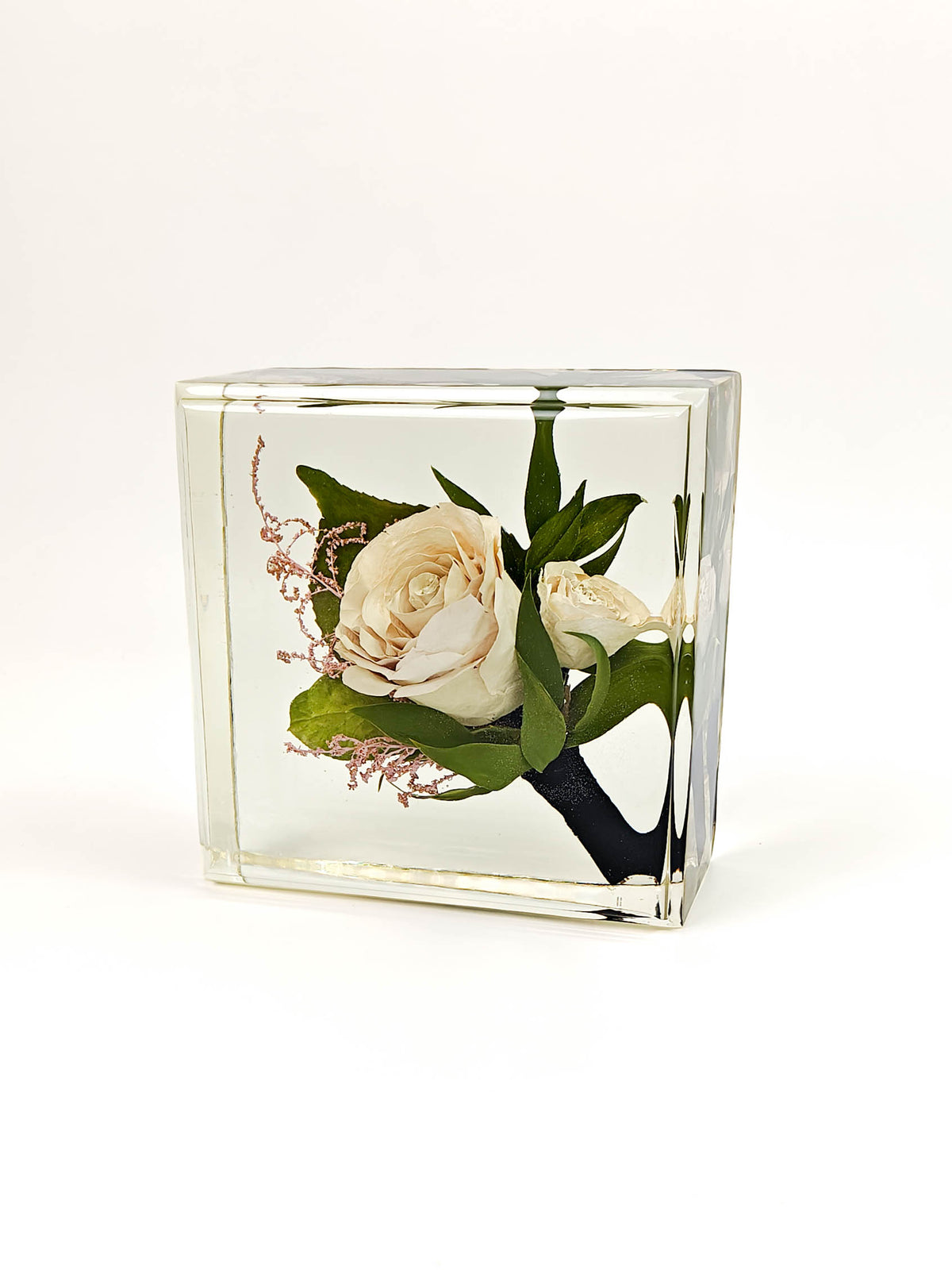 A square clear glass keepsake block containing a preserved floral arrangement with cream roses, green leaves, and delicate pink sprigs, photographed on a clean white background.